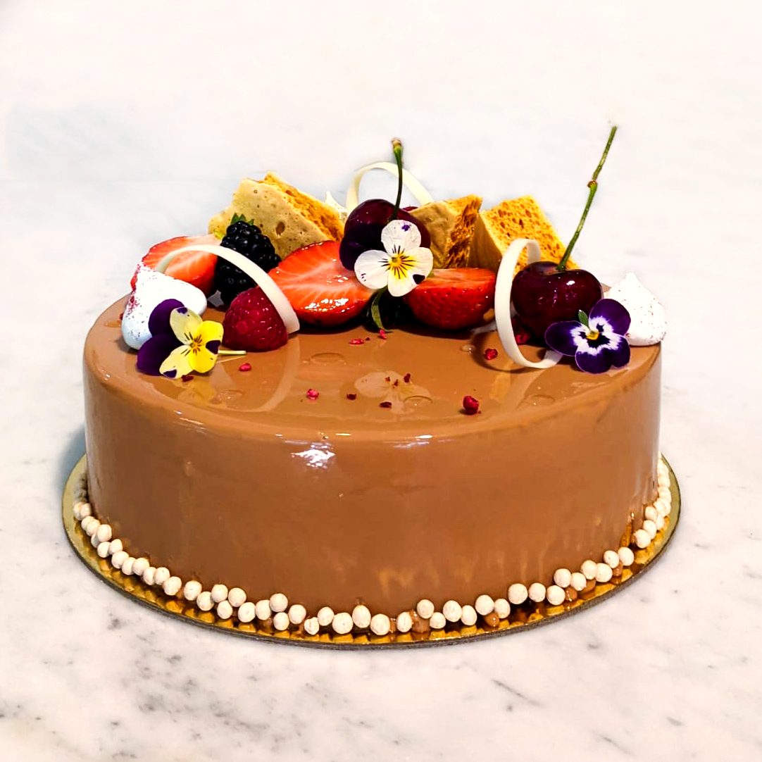 A glossy chocolate and salted caramel mousse cake from Strawberry Fields Patisserie, decorated with fresh berries, cherries and edible flowers